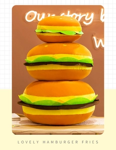 Three sizes of plush hamburger pillows stacked on a wooden table with warm lighting