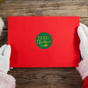 Green Merry Christmas gold foil sticker on a red envelope held by Santa gloves