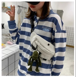 Detailed shot of white canvas chest bag featuring a green dinosaur plush charm on striped shirt