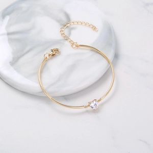 Polished gold heart bracelet with a sparkling white zircon on a white marble background