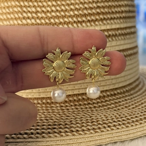 Golden sunflower earrings with white pearl drops held against a summer straw hat background