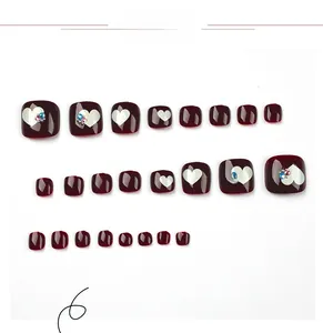 Detail of burgundy press-on toenails featuring white heart patterns and colorful rhinestones