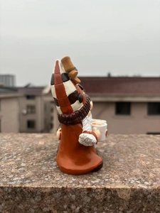 Side view of a resin gnome figurine wearing a brown plaid hat holding two coffee cups