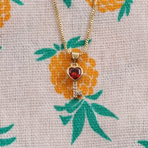 Detail of a gold-plated key necklace featuring a red heart crystal and pave accents