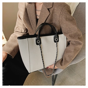 Detail of a large beige tote bag with black leather trim and decorative metal chain strap