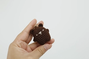 Close-up of a tiny handmade brown crochet bear keychain held by hand against white background