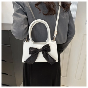 White shoulder bag with large black bow detail shown on a model in a minimalist room