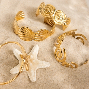 Detailed shot of 18K gold plated ocean-themed bangles with shell and wave patterns on sand