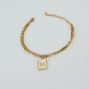 Detalle de la pulsera dorada de doble cadena tipo clip con un colgante cuadrado de letra M