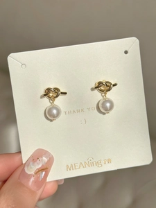 Macro detail of gold knot stud earrings with white pearls on a minimalist brand card