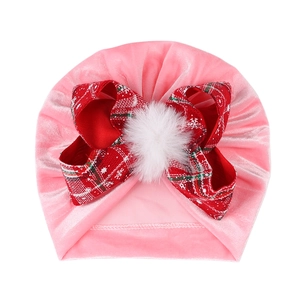 Pink velvet baby turban hat with a red plaid bow and white faux fur pompom detail