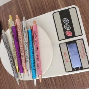 Set of 7 colorful glitter ballpoint pens featuring funny weekday themed phrases on a scale