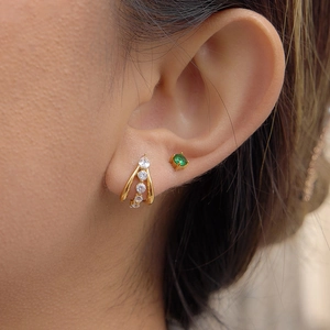 Gold C-shape earrings with clear zircons and a green gemstone stud shown on an ear