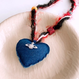 Dettaglio di un ciondolo a cuore in denim con logo saturno argento e cordino intrecciato