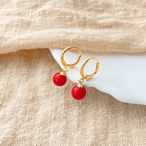 Close-up of gold hoop earrings with round red pearl pendants on a white ceramic tray