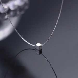 Close-up of a silver cube pendant on a fine box chain reflecting on a dark glossy surface
