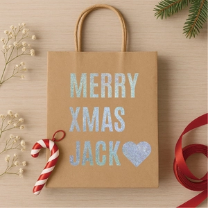 Brown kraft paper gift bag with glitter Merry Xmas Jack text on light wood background
