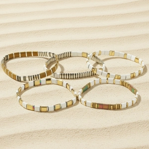 Close-up of five Bohemian Tila bead bracelets layered on beach sand with golden finish