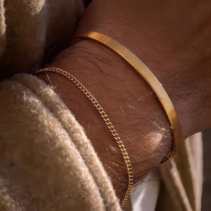 Detail of golden C-shaped cuff and Cuban chain bracelet on wrist with beige sleeve