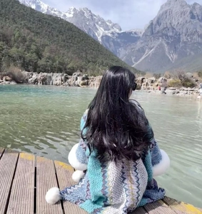 Detailed back view of a blue-green wavy knit shawl with white pom-pom trim against mountains