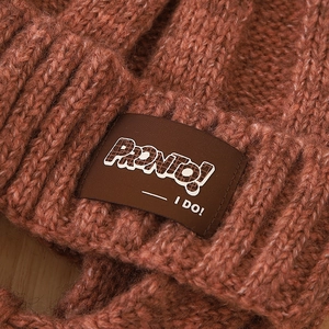 Detail of red-brown chunky knit hat featuring a sewn patch with Pronto and I Do text