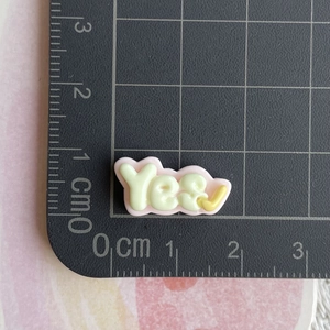 Close-up of a mini yellow and pink resin 'yes' word charm on a black measuring mat