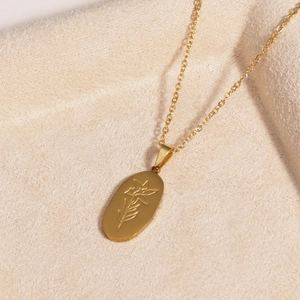 Detailed close-up of golden stainless steel oval pendant with floral engraving on beige fabric