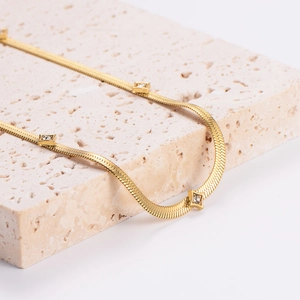 Gold flat snake chain necklace with square zircon accents on a beige stone surface