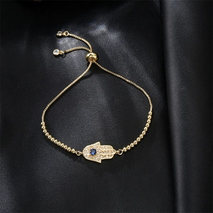 Golden Hamsa hand bracelet with zircon pavé and blue evil eye on black leather background