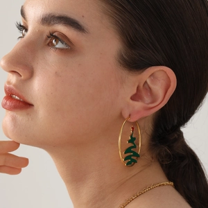 Golden hoop earrings with green Christmas tree charms worn by a model on a beige background