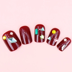 Glossy burgundy press-on nails with assorted colorful rhinestones on pink background