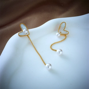 Asymmetric heart and pearl dangle earrings in gold finish on white and brown background