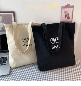 Detalle de bolsos de lona en crema y negro con diseño de ojos y estampado de texto sh!