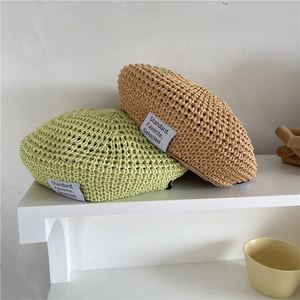 Light green and khaki woven straw beret hats displayed on a minimalist white platform