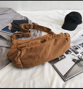 Brown canvas crossbody bag displayed on white bedding with lifestyle magazines and a cap