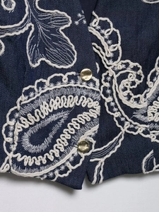 Close-up of dark blue denim vest featuring intricate beige embroidery and vintage metal buttons