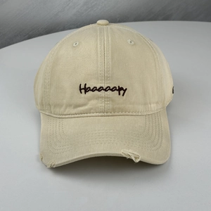 Detail of embroidered Haaaaapy letters and distressed ripped brim on beige baseball cap