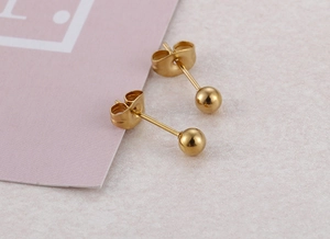 Close-up of golden ball stud earrings with butterfly backs on a textured beige surface