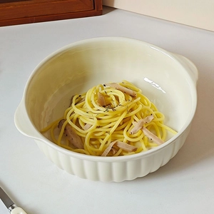 Cream ceramic baking bowl with handles holding pasta and herbs on a white surface