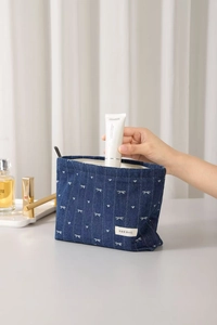 Detail of blue denim cosmetic bag with bow embroidery and a hand placing skincare inside