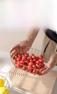 Detail showing hand holding gold wire fruit basket with red tomatoes on wooden tray