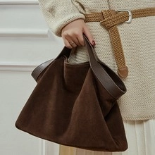 Close-up of a brown suede tote bag carried by a model against a white wall