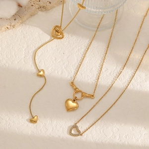 Trois colliers cœur plaqués or 18 carats sur surface beige texturée avec lumière douce