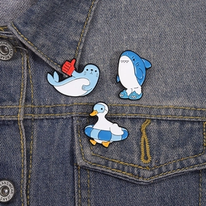 Three cartoon enamel pins featuring a seal, shark suit character, and duck on denim jacket