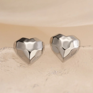 Polished silver heart-shaped stud earrings with geometric facets on a textured surface