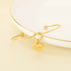 Detail eines goldenen Armreifs mit Love-Herzanhänger auf einer weißen Keramikplatte