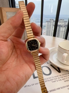 Detail of a square black dial watch with golden chain band held against a cafe backdrop
