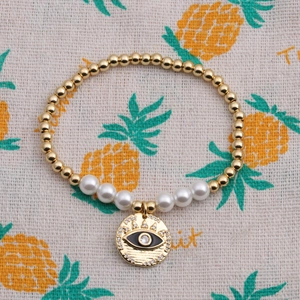 Golden bead and white pearl bracelet featuring an evil eye pendant on a fabric background