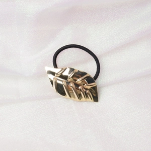 Detailed macro view of a golden leaf metal hair tie with a black elastic band on white fabric