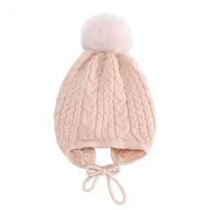 Detailed knit texture of beige baby winter hat featuring earflaps and a fluffy faux fur pom